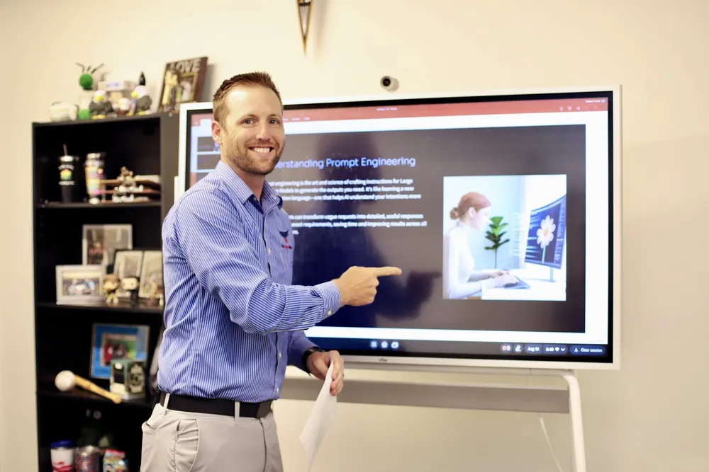 Man in a striped shirt pointing at a presentation slide on a digital screen in a room with a bookshelf of framed photos.
