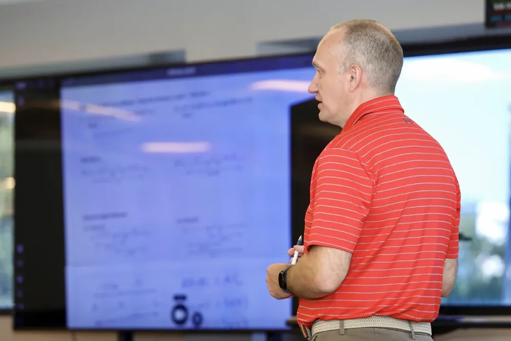 A man in a red striped polo shirt holding a pen, standing in front of a large screen displaying text during a presentation.
